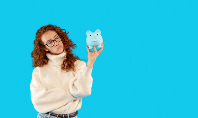 A young woman smiles while holding a cheerful blue piggy bank against a vibrant turquoise background