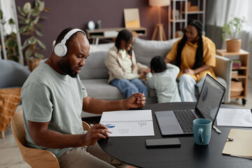 Fototapeta premium Man wearing headphones concentrating on work with laptop while family gathering in background interacting and playing with child in a cozy living room setting