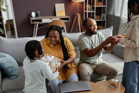 Family sharing joyful moment during special occasion in living room setting. Child presenting gift to mother while father observing with smile. Cozy, warm atmosphere capturing happiness