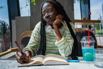 Young black woman is thoughtfully taking notes in her notebook while sitting at a table at a cafe. She is wearing a green and white striped shirt and has headphones around her neck