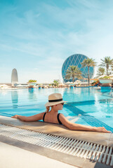 Girl in hat is in pool, leaning on side. Young woman with glasses and hat on vacation is relaxing...