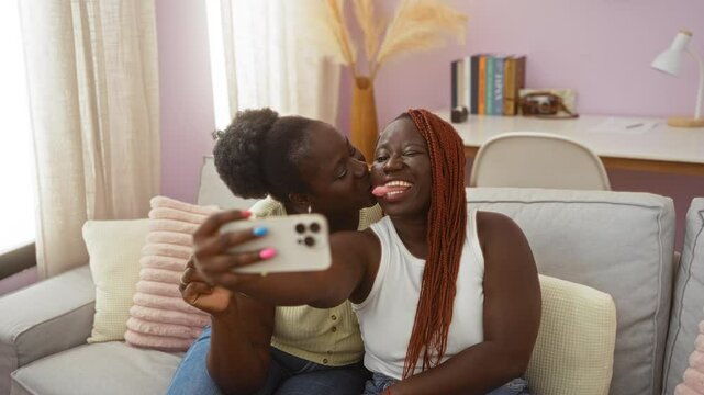 Two women taking selfies in a cozy living room, displaying love and happiness while sitting on a couch with pillows and books in the background