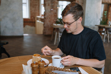 Man is sitting at a table in a cafe, enjoying a slice of layered cake and a cup of coffee. He is taking a bite of the cake with a fork