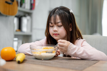 A young, adorable Asian girl is eating a bowl of cereal at the dining table for breakfast.