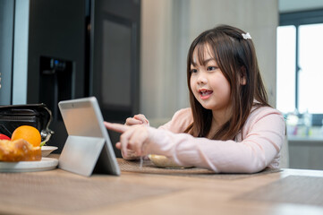 A young Asian girl is enjoying watching a cartoon on her digital tablet while having breakfast.