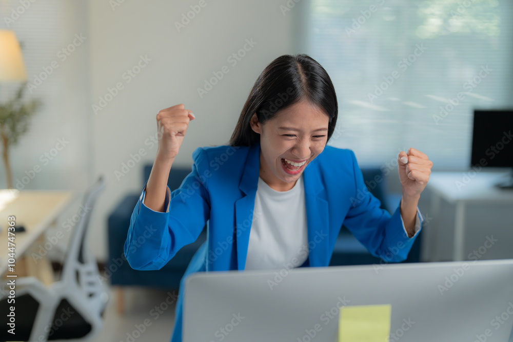 Wall mural young businesswoman is sitting at her desk with her fists raised in the air, she is happy with her w