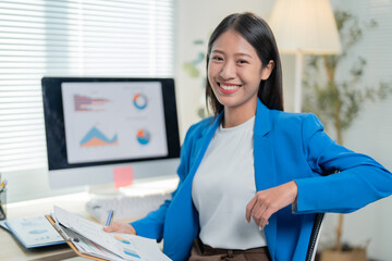 Asian businesswoman in a blue blazer reviews financial reports at her desk in a modern office, exuding confidence and success