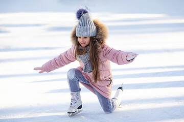 Cute little girl in warm pink coat, grey knitted hat and scarf, snood learning to skate on ice skating rink in park. Fall down and have fun. Winter sport, outdoors. Holiday and seasonal concept.