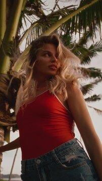 Vertical portrait: beautiful sexy girl stands next to a palm tree. Sporty fit blonde woman in a red swimsuit enjoys a tropical island on a summer vacation. Flirting look