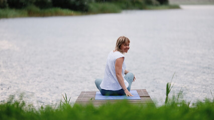 Fototapeta premium Young Caucasian woman sitting in lotus position on bridge by lake during rain. Woman enjoying fresh air. Outdoor meditation