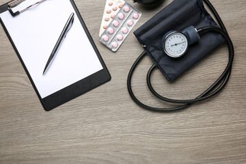 Blood pressure measuring device, pills and clipboard on wooden table, top view