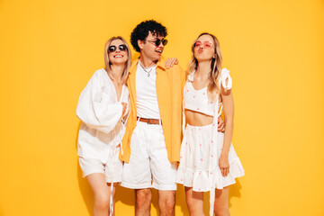 Fototapeta premium Group of young three stylish friends posing in studio. Fashion man and two cute female dressed in casual summer clothes. Smiling models having fun. Cheerful women and guy near yellow wall, sunglasses