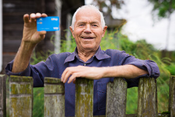 Happy elderly man showing credit card while standing by the fence of country house