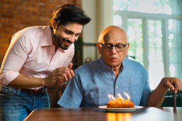 Indian son celebrating his elderly father’s birthday with cake, candle at home, joyful moments