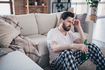 Photo of sad upset moody gloomy man wear sleepwear sitting on floor thinking indoors flat apartment