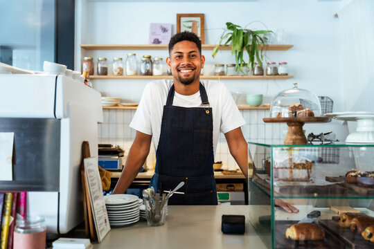 Bakery, happy portrait of hispanic black man in cafe ready for serving pastry, coffee and baked foods. Restaurant, coffee shop and confident waiter barista by counter for service, help and welcome