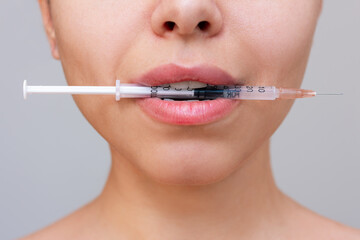Close up of a young caucasian woman's face with perfect lips with a syringe in the mouth on a grey...