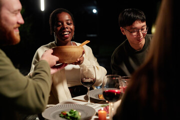 Group of smiling adult friends enjoying meal together outdoors focus on African American woman serving salad