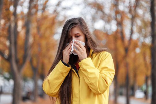 young woman stands park, dressed yellow jacket holding tissue to her nose as she sneezes. concept: seasonal cold rhinitis nose leaking use of paper handkerchiefs. influenza and arvi epidemics 
