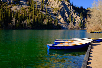 Naklejka premium Blue rowing boats moored at mountain lake shore with rocky cliffs and evergreen forest backdrop. Kolsai lake, Kazakhstan.