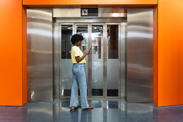 Happy afro woman using cell phone while waiting for elevator in hotel lobby