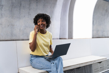 South African woman working using computer laptop talking on mobile phone at modern subway station