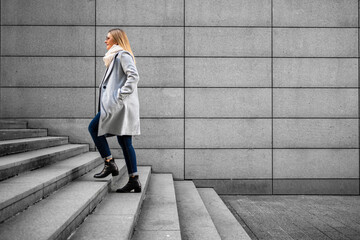 Middle-aged blonde woman in gray coat, scarf, jeans and sunglasses walking up concrete stairs  against background of concrete slabs in city on autumn day. Businesswoman. Side view.