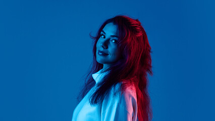 Side view portrait of young woman wearing white sweater, posing with little calm smile against blue studio background in neon light. Concept of human emotions, fashion, lifestyle, facial expression