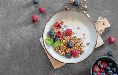 Granola with greek yogurt, raspberries and blueberries in a bowl on a wooden board on a dark background with fresh berries.