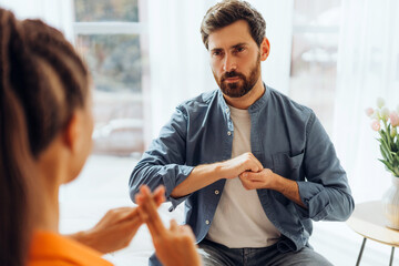 Pensive man and Latin woman, translator, sitting on sofa at home, speaking with sign language
