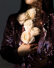 A woman in a shimmering blazer holds delicate white flowers against a backdrop of soft, dappled light, creating a moment of elegance