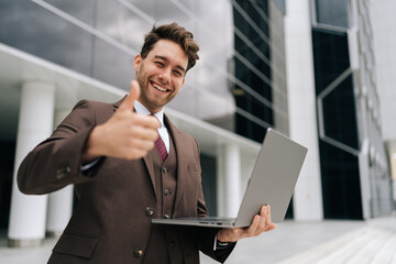 Portrait of cheerful successful businessman in urban setting, holding laptop and showing thumbs up gesture, smiling looking at camera, embracing confidence and professionalism in corporate world.