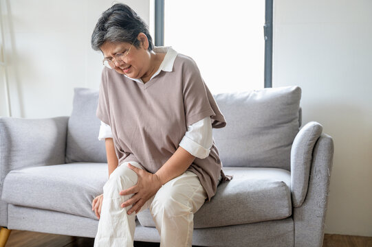 Mature Asian female suffering from knee pain osteoarthritis. Close up senior woman holding her knee due to physical injury while sitting alone at home. rheumatoid arthritis disease in Adult.