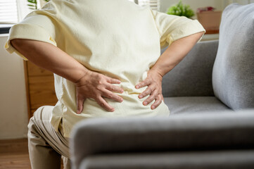 Fototapeta premium Senior Asian woman using hand pushing on her back because feeling back pain. An elderly person is sitting on a couch, holding their lower back in pain. highlighting discomfort and the need for medical