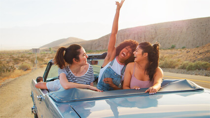Three Female Friends In Back Seat Of Classic Open Top Car On Road Trip Through Countryside