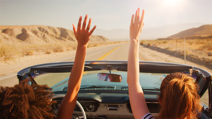 Two Female Friends Driving Classic Open Top Car On Road Trip In Countryside With Hands In The Air