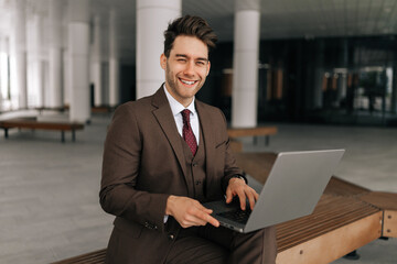 Fototapeta premium Portrait of positive successful businessman in suit sitting on urban bench by office building, working typing on laptop, looking at camera with happy expression. Concept of business communication.