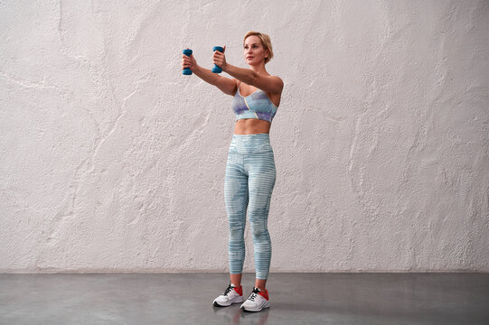 Female trainer in standing position feet shoulder-width apart arms straight arms stretched forward with dumbbells. Woman being in good physical shape has regular fitness training wears sportsclothes.