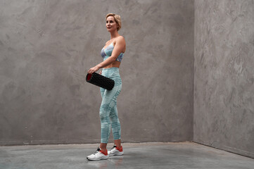 Side view of a young blonde woman in gym clothes after a workout, holding a workout roll. Young woman is working out in studio. It shows dedication and artistic expression.