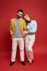 A middle aged couple enjoys a playful moment in fashionable attire on a striking red backdrop.