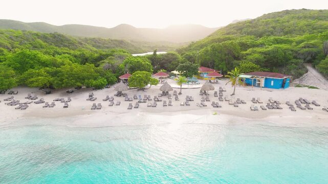Aerial pan of Kenepa Grande in Curacao, highlighting turquoise waters, sandy beach, and vibrant island greenery as sunrise light glistens on green lush mountains