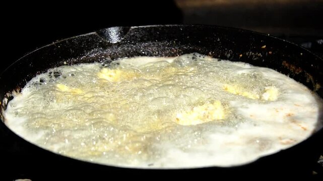 Close up while catfish nuggets or fish battered in breadcrumbs being placing in cast iron skillet or pan full of hot oil while deep frying over firepit or camp fire
