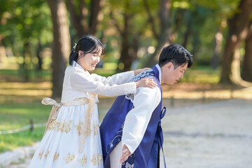 A Korean couple, a man in his 30s and a woman in her 20s, are wearing Hanbok and hugging each other in a park surrounded by green trees in Seoul, South Korea.