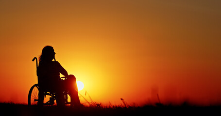 Obraz premium Silhouette of a woman in a wheelchair against an orange sky at sunset, thoughtfully gazing into the distance.