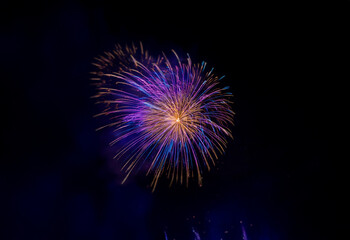 A single firework bursts in the night sky, exploding into a circular shape with a fiery center surrounded by vibrant purple and blue trails.