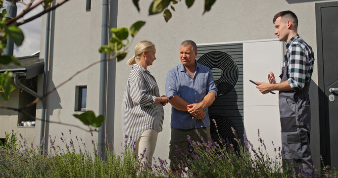 A technician discusses heat pump maintenance with a couple, using a tablet to illustrate points beside the unit.