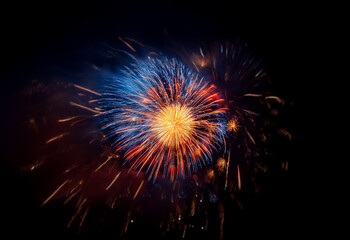 Obraz premium A single burst of fireworks explodes in the night sky, isolated against a black background. The fireworks are a mix of red, orange, and blue, creating a dazzling display.