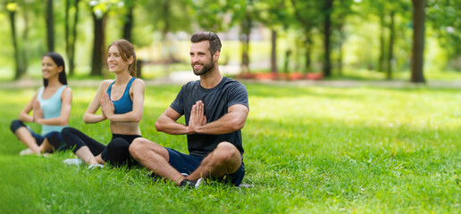 Group of people practicing yoga meditation on green grass summer park. Happy man and women in lotus pose, doing yoga exercises meditating. Wide panoramic banner image. Lotus pose. Selective focus.