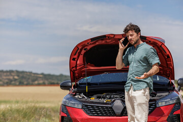 Young overwhelmed and angry man on vacation with reflective vest and broken car on the shoulder calling insurance on the phone, requesting a tow truck.