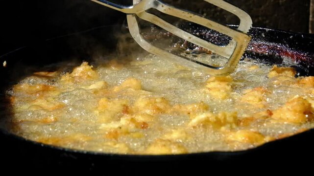 Super close up and slow motion while using tongs to stir catfish nuggets or fish in batter and breadcrumbs deep frying in cast iron skillet or pan over firepit while camping outdoors